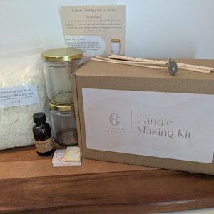 May include: A candle making kit in a brown cardboard box labeled Candle Making Kit, including a 14 oz bag of premium soy wax, two glass jars with gold lids, a small amber bottle of fragrance oil, wooden wick centering tools, and instructions.