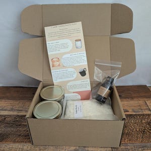 Pu&ograve; includere: Una scatola di cartone aperta contenente materiali per la produzione di candele. All'interno ci sono due barattoli di vetro con coperchi dorati, un sacchetto di cera, una bottiglia di olio profumato e le istruzioni. La scatola &egrave; su una superficie di legno.