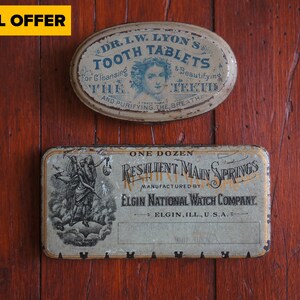 Puede incluir: Dos cajas de hojalata vintage. La caja superior es de forma ovalada y tiene el texto "DR. L.W. LYON'S TOOTH TABLETS" impreso en ella. La caja inferior es rectangular y tiene el texto "ONE DOZEN RESILIENT MAIN SPRINGS MANUFACTURED BY ELGIN NATIONAL WATCH COMPANY. ELGIN, ILL, U.S.A." impreso en ella.