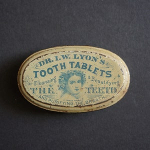 Puede incluir: Una lata ovalada antigua de pastillas dentales Dr. I.W. Lyon. La lata tiene un fondo color crema con texto azul y un retrato de mujer. El texto incluye "Tooth Tablets" y "For Cleansing The Teeth and Purifying the Breath."