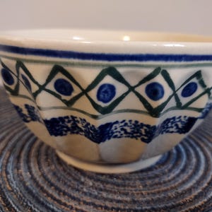 May include: A white ceramic bowl with a blue and green geometric pattern. The bowl has a scalloped edge and a blue band around the rim. The design includes blue dots within green diamond shapes.