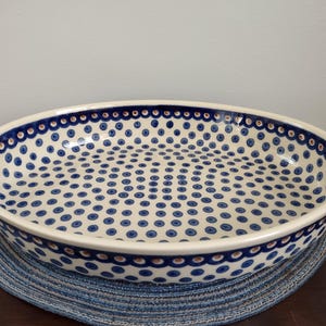 May include: Oval ceramic serving dish with a white base and a blue polka dot pattern. The rim is decorated with a scalloped blue border. The dish is placed on a blue woven placemat.