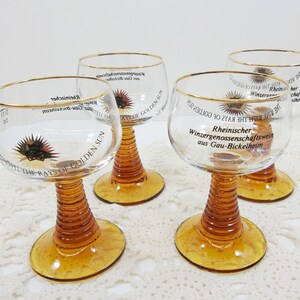 May include: Four clear glass wine glasses with amber stems and gold rims. Each glass has a different inscription on the side, including "At the Rays of Golden Sun" and "Rheinischer Winzergenossenschaftswein aus Gau-Bickelheim".