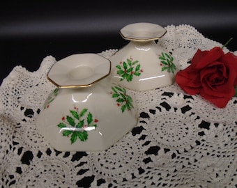 May include: Two white ceramic candle holders with a gold trim and a holly design. The holders are on a white crocheted doily.