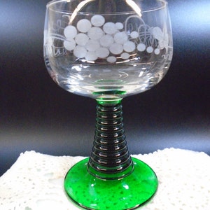 May include: A clear wine glass with a green base and a black, ribbed stem. The glass features a white grape design etched on the bowl. The base is a vibrant green, and the stem has a textured, dark appearance.