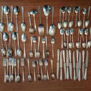May include: A large collection of silver-toned metal flatware including various sizes of spoons, forks, and knives. The set features multiple dinner forks, salad forks, soup spoons, teaspoons, and dinner knives arranged in a grid-like pattern.