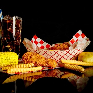May include: A red and white checkered paper takeout container with fried chicken, a yellow corn on the cob, and curly fries. A glass of cola with ice and a blue and white striped straw is in the background.
