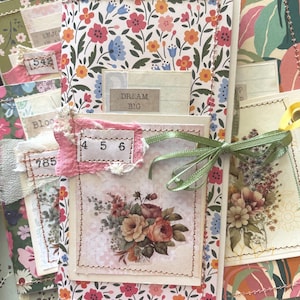 May include: A handmade paper journal featuring a floral pattern with red, orange, and blue flowers. The cover includes a layered collage with a tag reading "DREAM BIG," a pink fabric scrap with the numbers "4 5 6" stitched on, and a green ribbon tie.