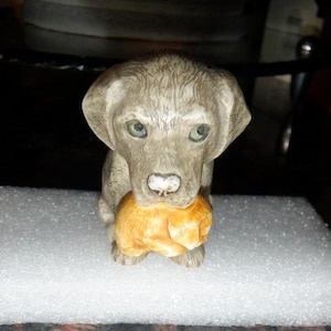 May include: A small gray ceramic figurine of a dog holding a brown and orange puppy in its mouth.