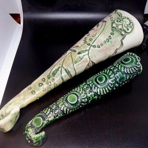 May include: Two ceramic decorative pieces. One is off-white with green floral and beaded designs. The other is dark green with circular patterns. Both have a curved shape and are placed on a dark surface.