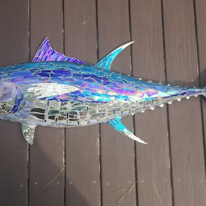 May include: A mosaic sculpture of a tuna fish, primarily blue with iridescent highlights. The fish is crafted from small, irregular pieces, creating a textured surface. The fins and tail are sharply pointed, and the fish is displayed against a wooden background.