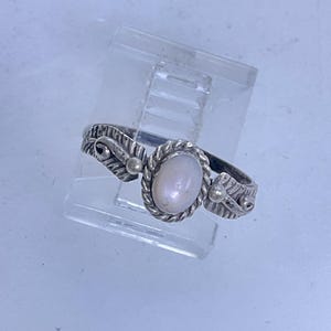 May include: A silver ring with an oval, light-colored stone set in a twisted rope design. The band features leaf and bead details on either side of the stone. The ring is displayed on a clear plastic stand.