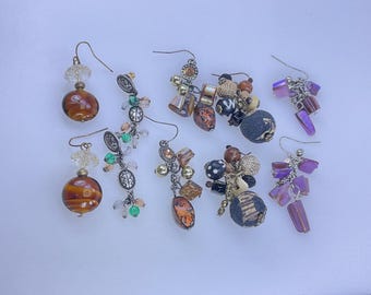 May include: A collection of handmade earrings with various designs. Some earrings feature amber-colored glass beads, while others have clusters of beads in brown, green, and purple hues. The earrings are displayed on a white surface.