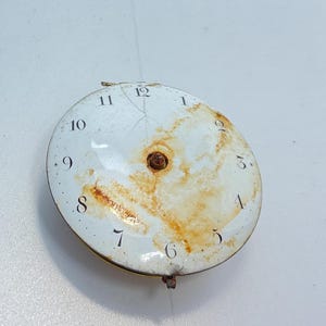 May include: A white clock face with black Roman numeral markings for the hours 1 through 12. The clock face is slightly damaged and has rust stains.