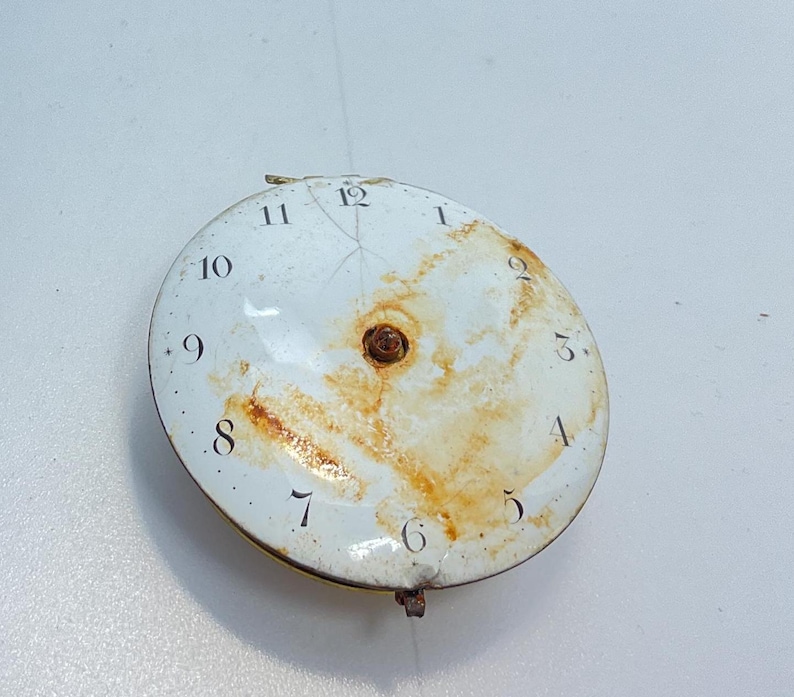 May include: A white clock face with black Roman numeral markings for the hours 1 through 12. The clock face is slightly damaged and has rust stains.