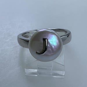 May include: A silver ring with a white pearl featuring the letter "J" in silver.
