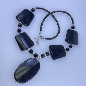 May include: A black necklace featuring a variety of shapes. The necklace has a central oval pendant and several rectangular and round beads. The beads are connected by a black beaded chain and silver-toned accents.