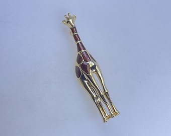 May include: A gold-toned giraffe brooch with a long neck and legs. The giraffe's body features brown spots and a brown neck. The brooch is a decorative accessory.