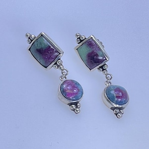 May include: A pair of silver earrings featuring rectangular and oval-shaped gemstones. The rectangular stones have a green and purple pattern, while the oval stones are purple. The earrings have a dangling design.