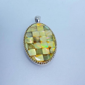 May include: Oval pendant with a checkerboard pattern of iridescent squares in shades of yellow, gold, and brown. The pendant has a silver-colored bail and a decorative, beaded border. The overall design is elegant and eye-catching.
