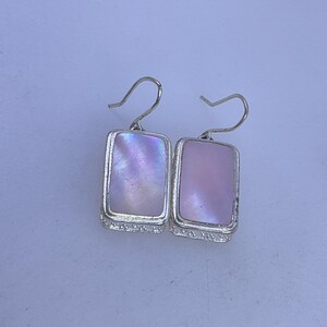 May include: A pair of rectangular earrings with iridescent pink and purple mother-of-pearl insets. The earrings have a silver-colored frame with a decorative edge and a hook closure.
