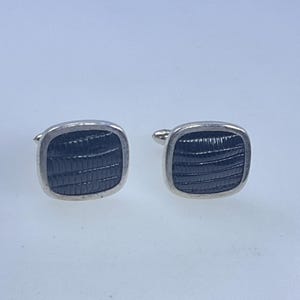 May include: A pair of square cufflinks with a silver-colored border and a textured, dark blue center. The cufflinks are displayed on a white surface, showcasing their design and details.