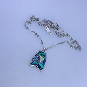 May include: A silver-toned necklace with a pendant featuring a yin-yang symbol. The pendant has a blue and green iridescent abalone shell design. The necklace is on a white background.