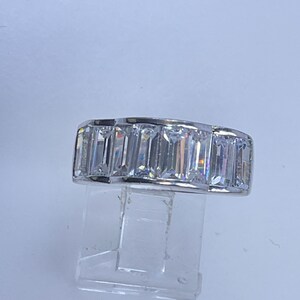 May include: A silver ring featuring a row of rectangular, clear gemstones. The gemstones are set closely together, creating a continuous band of sparkle. The ring is displayed on a clear stand against a white background.