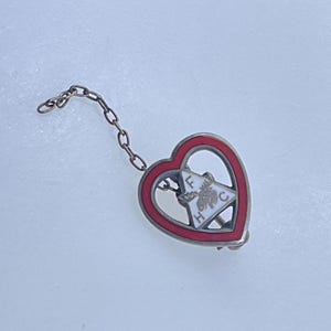 May include: Vintage heart-shaped pin with a red border and a small chain. The pin features a white emblem with the letters "FHC" and a decorative design. The pin is made of metal and has a clasp on the back.