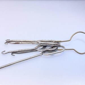 May include: A collection of vintage silver-toned knitting tools, including crochet hooks and a large loop tool. The tools are arranged on a white surface, showcasing their various shapes and sizes. The metal has a slightly aged appearance.