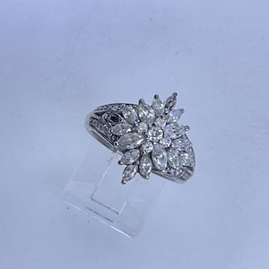 May include: A silver ring featuring a cluster of marquise-cut and round-cut clear gemstones, arranged in a floral design. The band has intricate detailing. The ring is displayed on a clear stand.