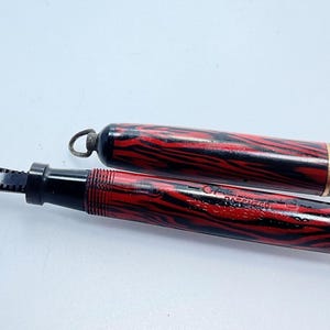 May include: A red and black striped fountain pen with a gold band and the text "The Conklin Crescent" and "C.A.E. Patented" on the barrel.