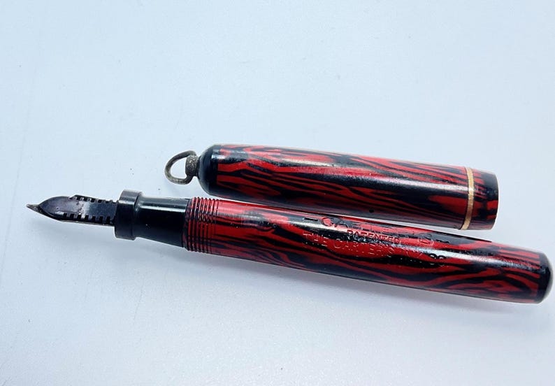 May include: A red and black striped fountain pen with a gold band and the text "The Conklin Crescent" and "C.A.E. Patented" on the barrel.