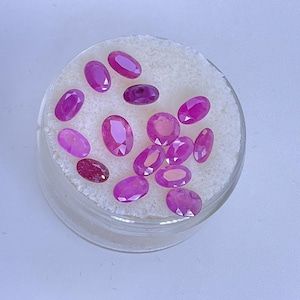 May include: A collection of oval-shaped, pink gemstones displayed in a clear, round container. The gemstones vary in size and shade, with a range of pink hues. The container sits on a white, textured surface.