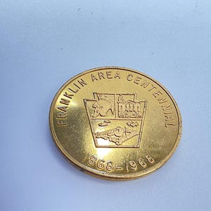 May include: A gold-colored coin with the text "FRANKLIN AREA CENTENNIAL" around the edge. The center features a crest with images and the dates "1868-1968".