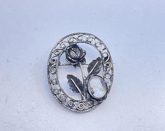 May include: Oval silver-tone brooch featuring a rose and cameo design. The brooch has an open filigree border with a rose at the top and a cameo on the right side. The cameo has a white profile against a darker background. The brooch is a vintage style.