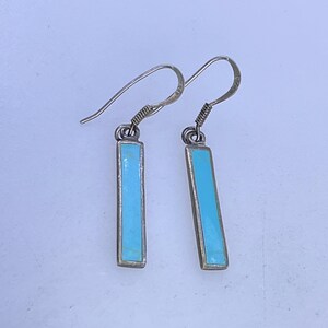 May include: A pair of silver-toned dangle earrings. Each earring features a rectangular, light blue stone set within a silver frame. The earrings have a simple, elegant design with a hook closure.