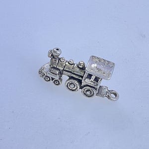 May include: A small, detailed silver-colored train charm. The charm features a locomotive with wheels, a smokestack, and a rectangular car. It has a loop for attaching to a bracelet or necklace. The charm is on a light blue background.
