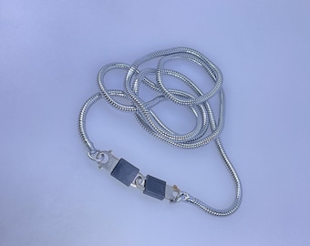 May include: A silver-colored necklace with a snake chain design. The necklace has a clasp with two black rectangular pieces. The chain is coiled on a light blue surface.