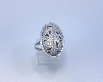 May include: An oval-shaped silver ring featuring an intricate openwork design of carved leaves and swirling patterns. The metal has a polished finish with darkened recessed areas that highlight the detailed floral-inspired engravings on the face.