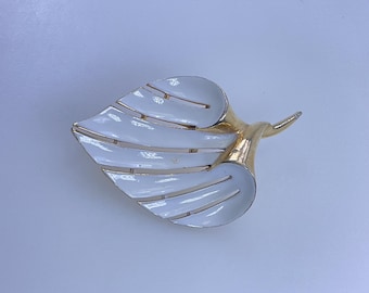 May include: A vintage leaf-shaped brooch with a white enamel finish and gold-tone metal accents. The brooch features a detailed design with multiple layers, creating a textured appearance. A classic accessory.