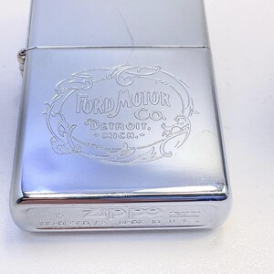 Puede incluir: Un encendedor Zippo plateado y brillante con el logotipo de Ford Motor Co. grabado en la parte delantera. El logotipo est&aacute; enmarcado en un &oacute;valo decorativo con las palabras "Detroit, Mich." debajo. La parte inferior del encendedor dice "Zippo" y "Made in USA".