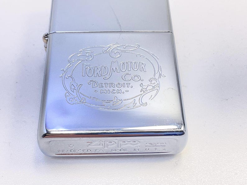 Puede incluir: Un encendedor Zippo plateado y brillante con el logotipo de Ford Motor Co. grabado en la parte delantera. El logotipo est&aacute; enmarcado en un &oacute;valo decorativo con las palabras "Detroit, Mich." debajo. La parte inferior del encendedor dice "Zippo" y "Made in USA".