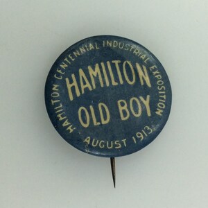 May include: A blue button with white text that reads "Hamilton Centennial Industrial Exposition Old Boy August 1913".