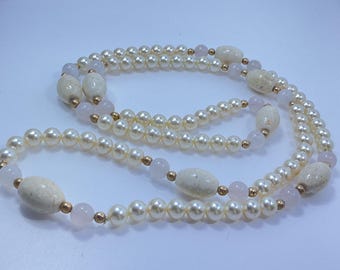 May include: A long necklace featuring multiple strands of faux pearls, interspersed with oval-shaped cream-colored beads and small pink beads. Gold-colored metal accents connect the beads. The necklace is displayed on a light background.