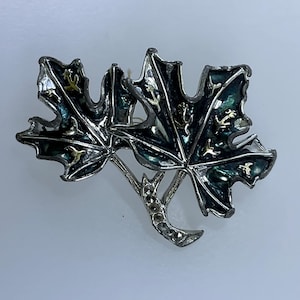 May include: A silver and black enamel brooch featuring two stylized maple leaves with gold accents. The leaves have a textured surface and are connected by a silver stem.