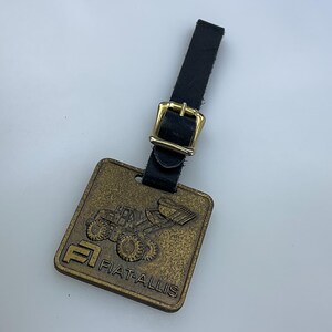 May include: A gold-colored metal keychain with a black leather strap and a gold buckle. The keychain features a small image of a Fiat-Allis tractor.