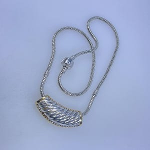 May include: Silver-toned necklace with a textured, curved pendant featuring gold accents. The necklace has a snake chain design and a clasp. The pendant is approximately 1.5 inches long, and the necklace is about 18 inches in length.