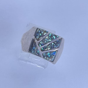 May include: A silver ring with a rectangular face. The face is inlaid with iridescent abalone shell pieces in a geometric pattern. The ring is displayed on a clear plastic stand against a light blue background.