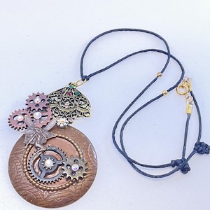 May include: A steampunk-style necklace featuring a large, round, copper-colored pendant adorned with gears, a fairy, and rhinestones. The pendant is suspended from a black cord with a gold clasp. The necklace has a unique, intricate design.